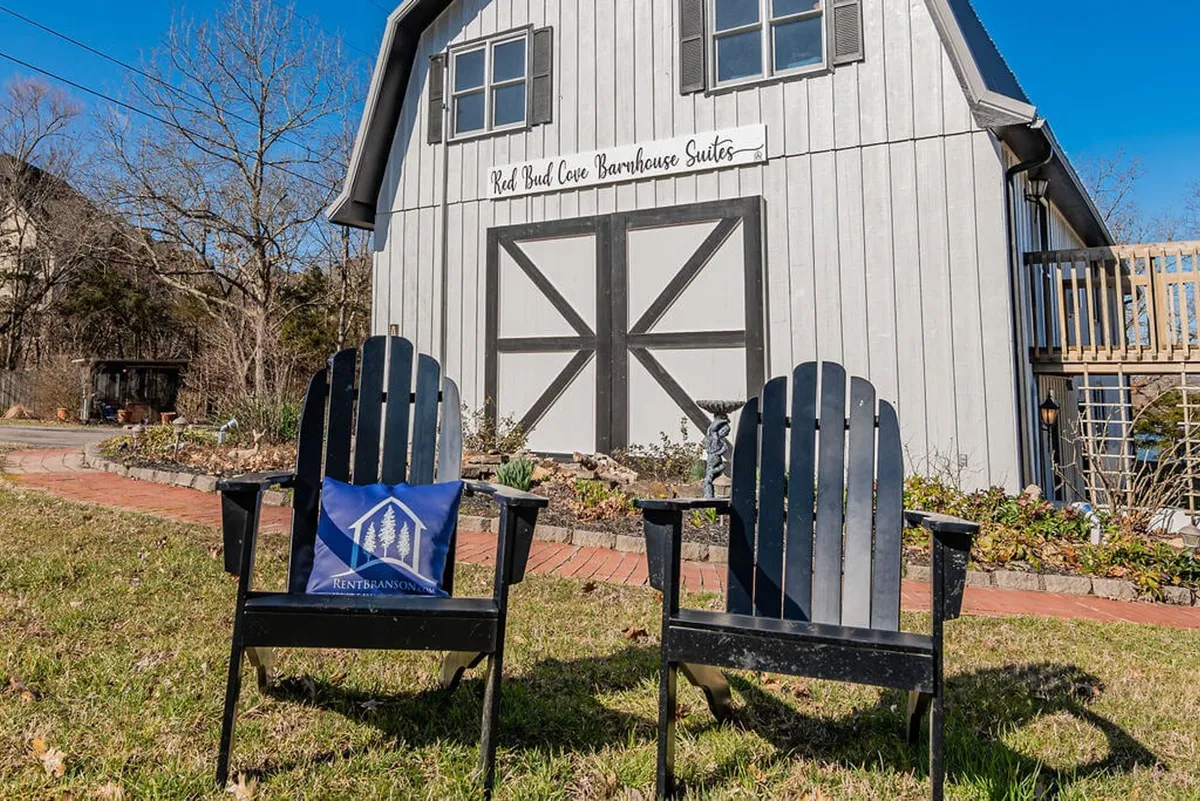 The image showcases a charming barn-style building nestled on the lakefront, with inviting Adirondack chairs arranged to face the water. A decorative pillow adds a cozy touch, and the golden light of sunset bathes the scene, suggesting an ideal spot for relaxation, conversation, or stargazing.