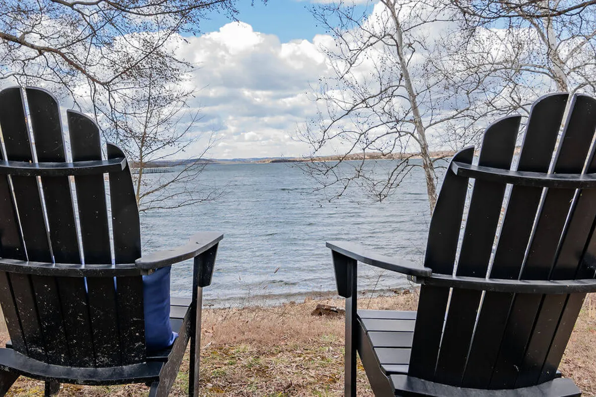 Two black Adirondack chairs are positioned at the edge of a grassy area, facing out over the tranquil lake. The sky is either early morning or evening, casting a soft glow over the calm water. The chairs suggest an inviting spot for two people to relax, take in the view, or share quiet moments together.