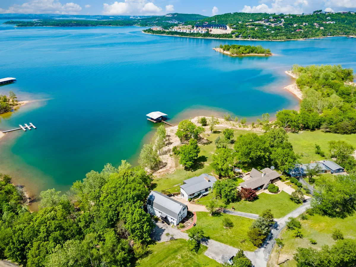 A serene lakeshore scene with blue water framed by lush green trees, several houses nestled along the shore, and a boat dock extending into the water. The setting is inviting and picturesque, evoking the appeal of lakeside activities like swimming, boating, or paddleboarding.