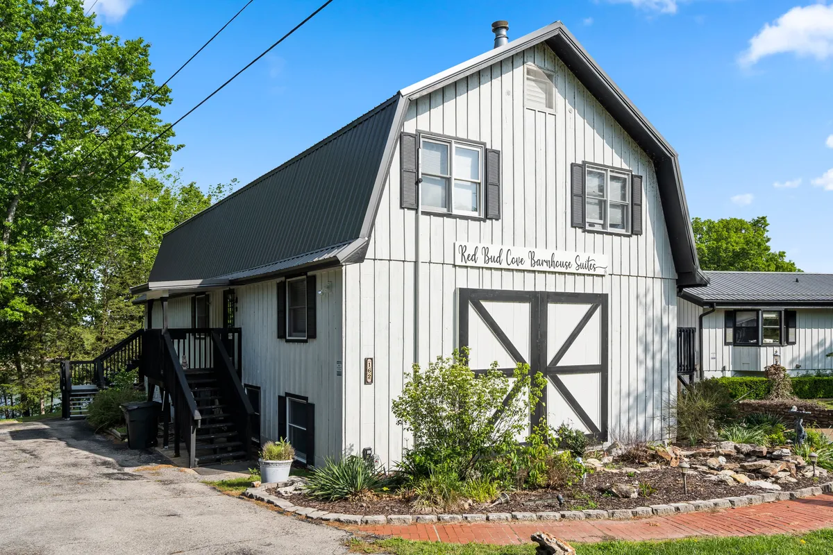 An inviting exterior shot of the barn-style suite, with prominent signage reading 'Red Bud Cove Barnhouse Suite' and a landscaped garden in the foreground. The porch and surrounding greenery project a welcoming and serene vibe.