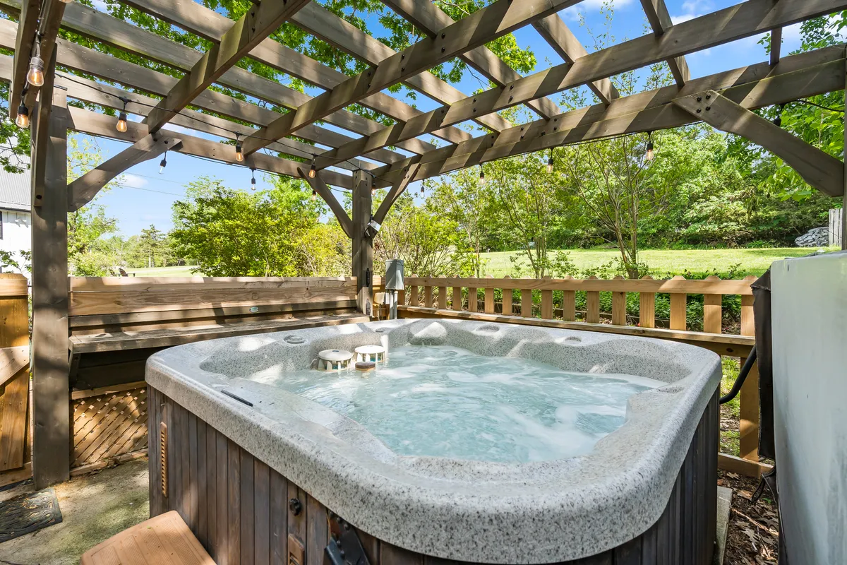A serene outdoor hot tub sits beneath a wooden pergola, surrounded by lush greenery. The inviting water steams gently, with comfortable seating nearby, creating an ideal spot for relaxation amidst nature.