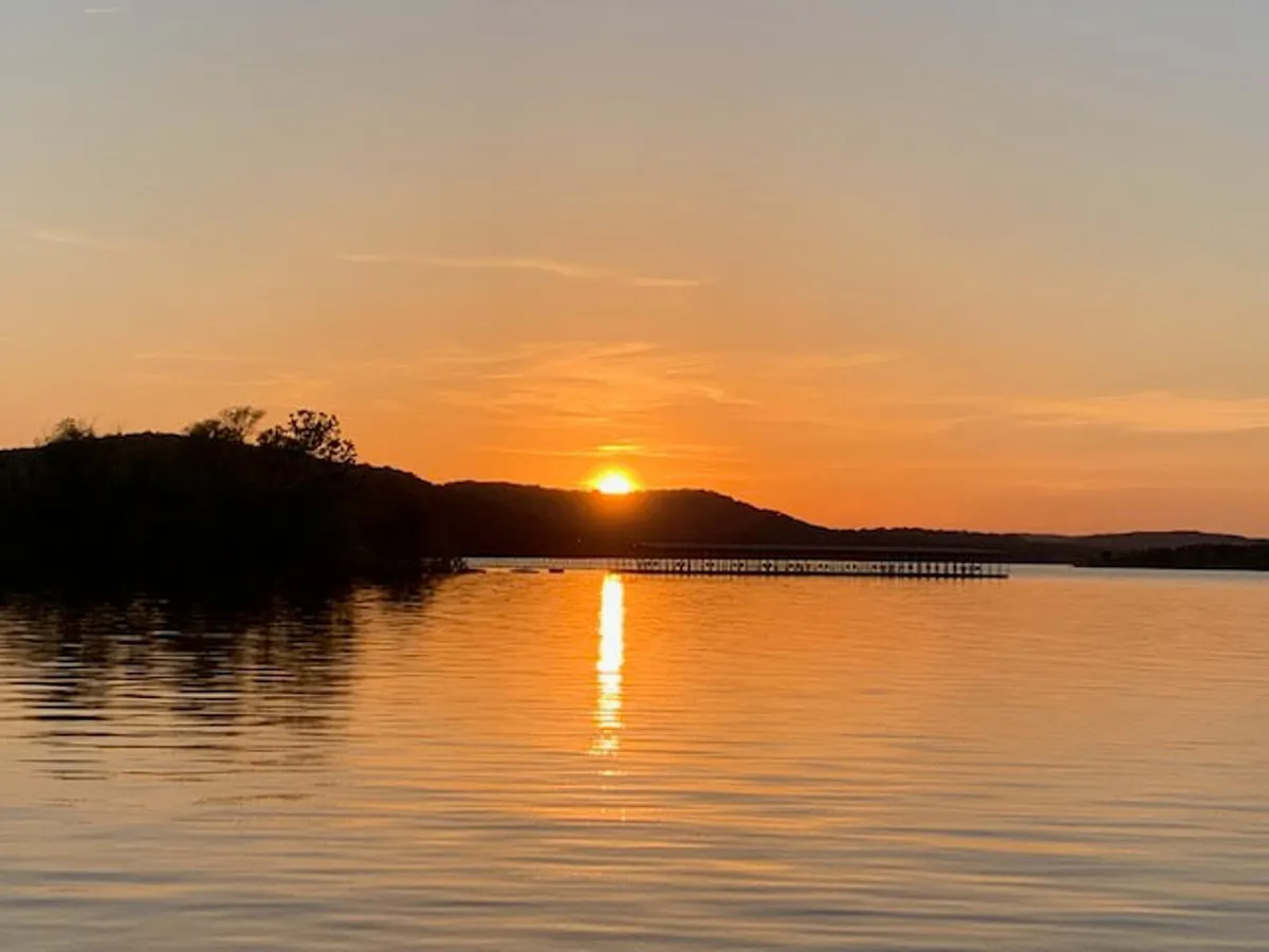 A picturesque sunset casts vibrant colors over the lake, with shimmering reflections on the water. A pier extends into the lake, and gentle hills rise in the distance. The scene is serene and visually stunning, capturing the magic of the lakeside at dusk.
