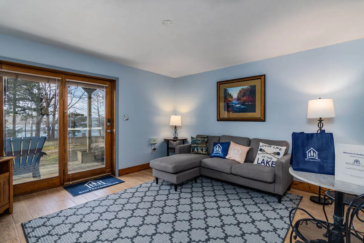 A comfortable living room with a plush sofa, tasteful artwork, a lamp casting a cozy glow, and a glass door that leads directly to the lakefront patio. The serene scene outside is hinted at through the doorway, blending indoor relaxation with outdoor allure.