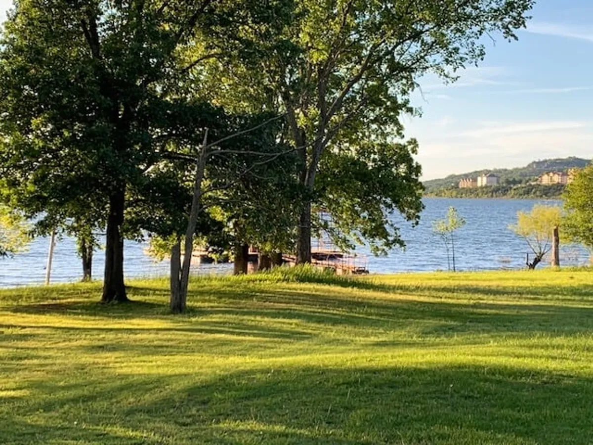 A peaceful exterior scene showing lush green grass, large shady trees, and calm lake waters with the distant shoreline. The space is set up for lakeside picnicking, fishing, and boating, and offers sweeping sunset views that appeal to families seeking both activity and relaxation.