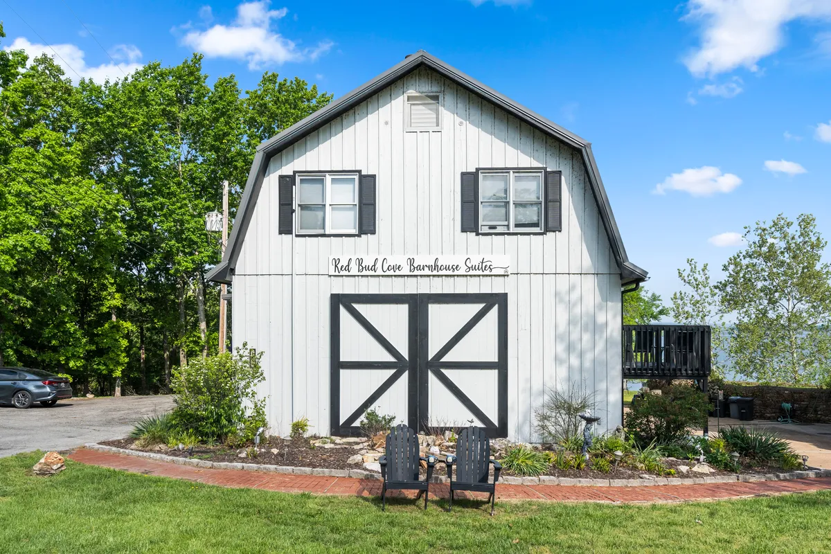 The barn-style exterior of a guest suite is featured, with black shutters, double doors, and signature signage reading 'Red Bud Cove Barnhouse Suites.' Two chairs sit invitingly at the entrance, set amidst a tranquil, green outdoor area.