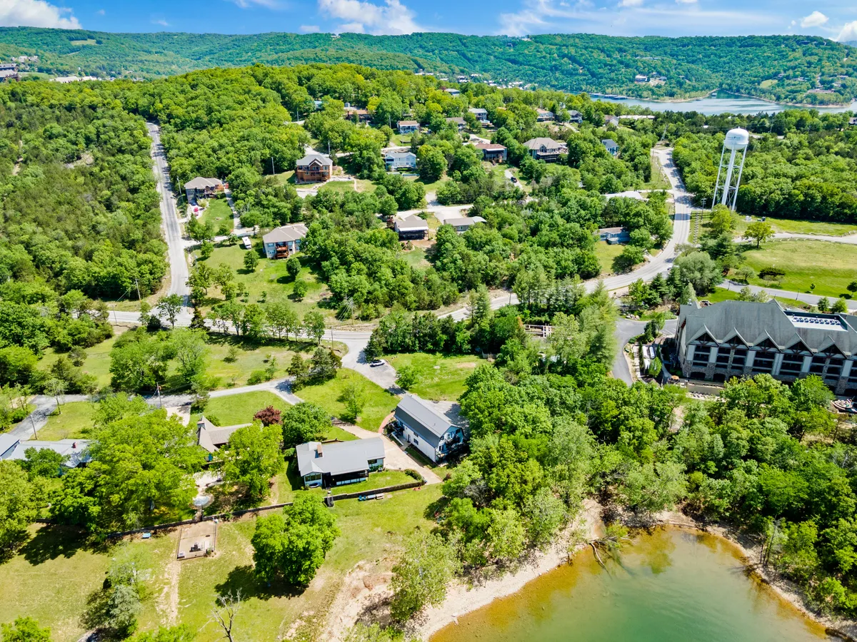 A panoramic view features lush greenery, a scenic waterway, nearby residential homes, and a distant water tower. The scene suggests access to both natural beauty and a vibrant local community, emphasizing the proximity to lakeside relaxation and outdoor activities such as hiking and boating.