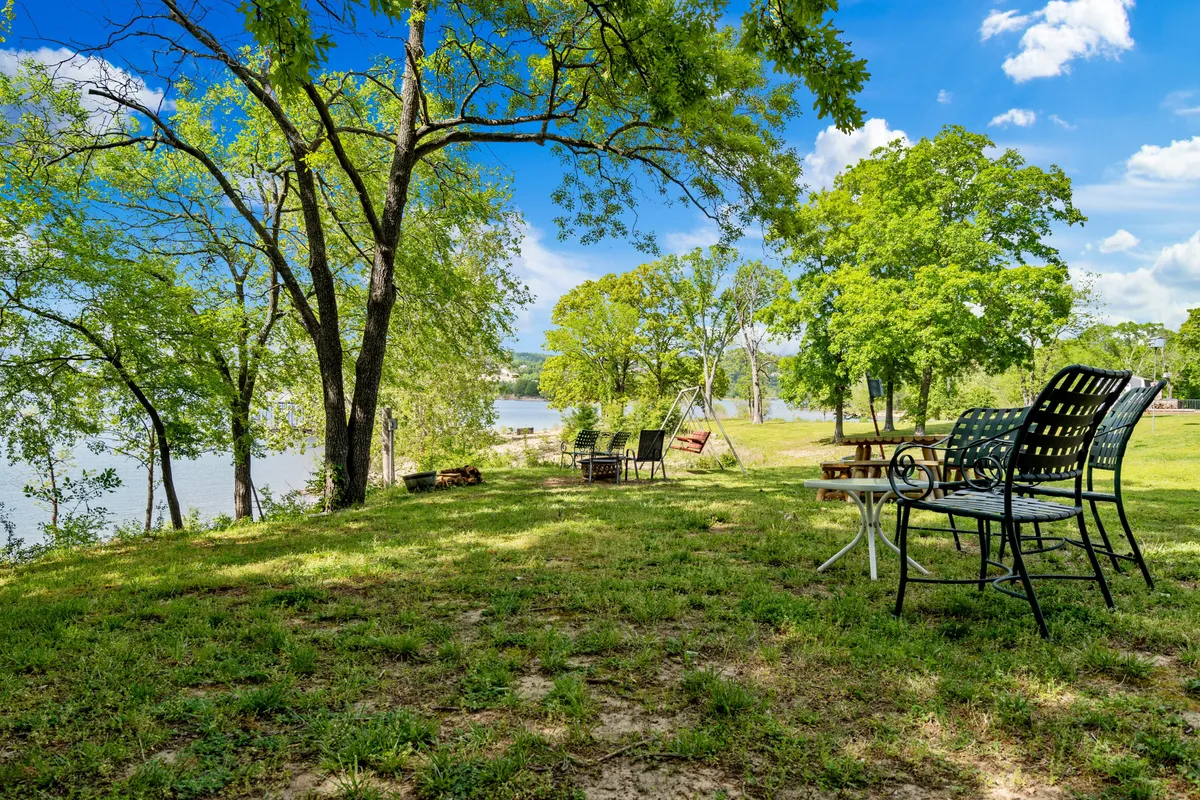 A grassy lakeside area features Adirondack chairs overlooking the water, shaded by trees, with a swing set nearby. The scene is set for picnics, barbecues, and playful gatherings, with a beautiful water view.